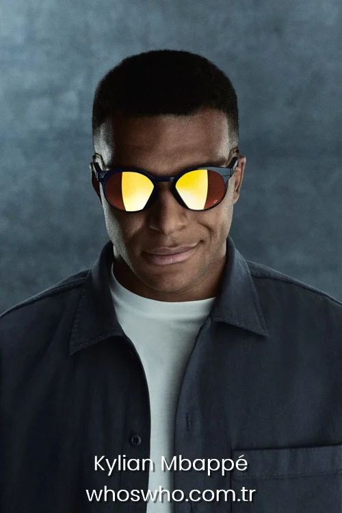 Close-up portrait of Kylian Mbappé smiling slightly while wearing dark round Oakley sunglasses with bright orange reflective lenses, styled in a dark grey jacket over a white t-shirt.