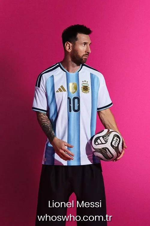 Argentine forward Lionel Messi holding a football and looking sideways while posing in the official white and light blue Argentina national team jersey against a bright pink background.