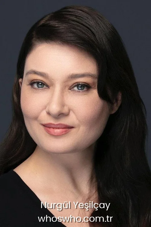 Close-up headshot portrait of smiling Turkish actress Nurgul Yesilcay with long dark hair, wearing a black top against a dark gray background.