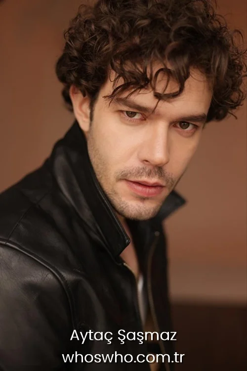Close-up headshot portrait of handsome Turkish actor Aytac Sasmaz looking intensely at the camera, with messy curly dark hair and stubble, wearing a black leather jacket against a soft brown background.