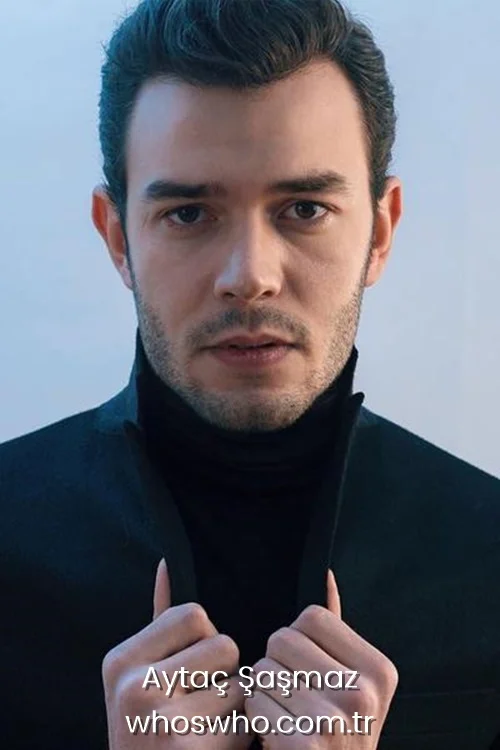 Close-up studio portrait of handsome Turkish actor Aytac Sasmaz looking directly at the camera with an intense expression, wearing a black turtleneck shirt and a high-collared black jacket against a light background.