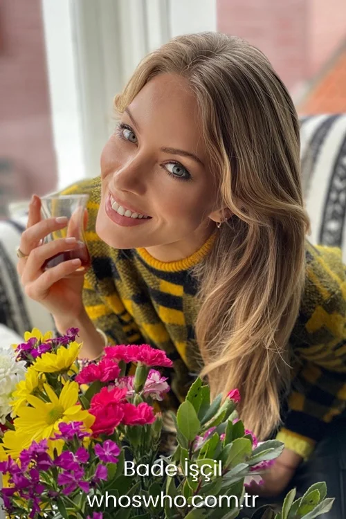 Turkish actress Bade Iscil smiling and holding a traditional Turkish tea glass behind colorful flowers