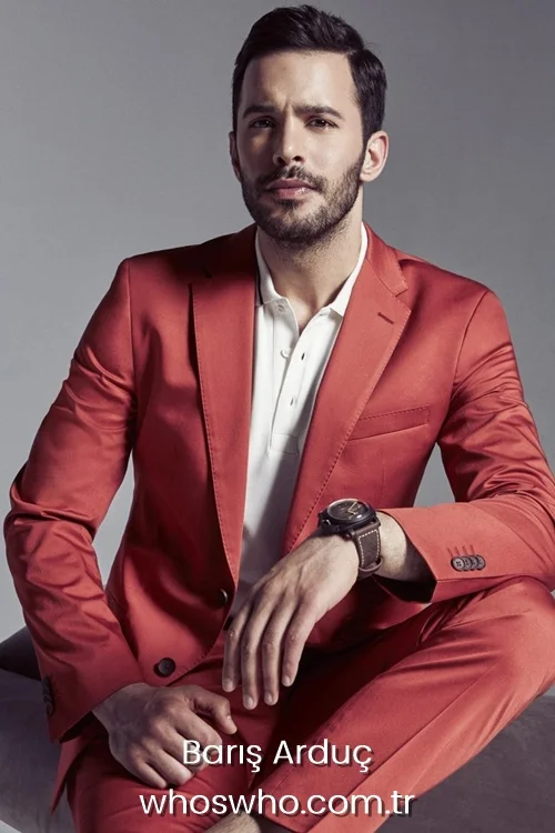 Handsome Turkish actor Baris Arduc posing with a serious expression, wearing a stylish terracotta red suit jacket and trousers over a white polo shirt, accessorized with a dark leather strap wristwatch.