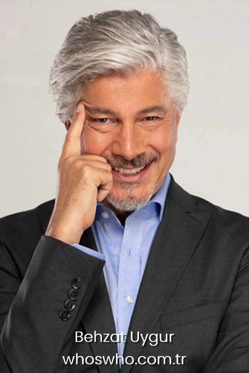 Smiling Turkish actor and TV presenter Behzat Uygur with grey hair, wearing a black blazer and blue shirt, posing with his finger on his temple.