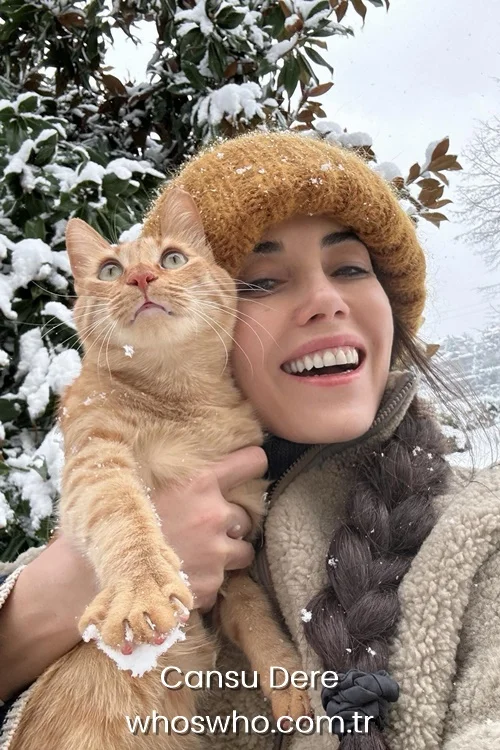 Cansu Dere smiling in the snow holding an orange tabby cat while wearing a yellow beanie.