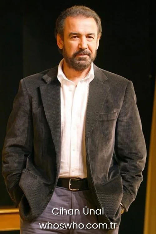Legendary Turkish actor Cihan Unal with a beard, wearing a dark grey corduroy blazer over an open-collar white shirt and grey trousers, posing with his hands in his pockets.