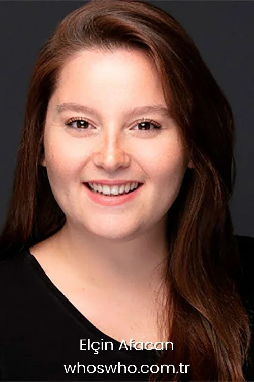 Close-up portrait of Turkish actress Elcin Afacan smiling broadly, showcasing her long brown hair and wearing a black t-shirt against a dark background.