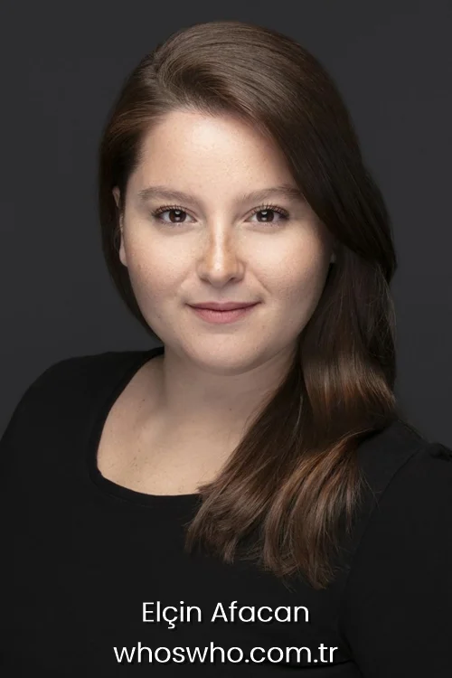 Close-up headshot of Turkish actress Elcin Afacan with long brown hair parted to the side, smiling gently and wearing a black scoop neck top against a plain dark grey background.