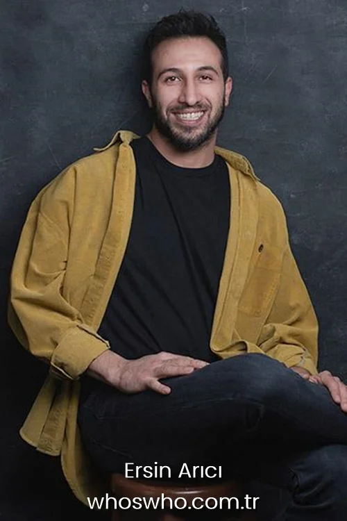 Smiling Turkish actor Ersin Arici with a beard, wearing an open mustard yellow corduroy shirt over a black t-shirt and dark jeans, sitting against a dark textured background.