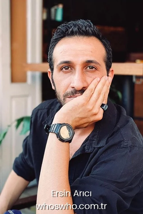 Close-up portrait of Turkish actor Ersin Arici with dark hair and short stubble, wearing a black shirt and a black wrist watch, resting his cheek against his hand.