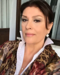 Recent close-up portrait of legendary Turkish actress Gulsen Bubikoglu looking elegant, wearing a white collared shirt under a leopard and floral print wrap, accessorized with a large pearl earring.