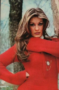 Classic portrait of Turkish actress Gulsen Bubikoglu with voluminous highlighted brown hair, wearing a vibrant red knit sweater and a long medallion necklace, posing outdoors next to a tree.