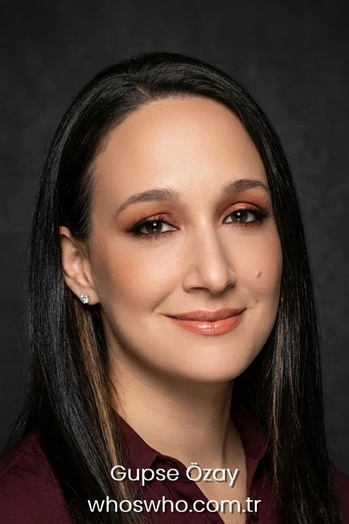 Close-up headshot portrait of Turkish actress and director Gupse Ozay smiling subtly, with long dark straight hair and warm makeup, wearing a burgundy collared shirt against a plain dark grey background.