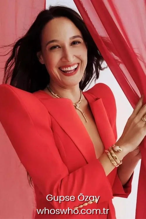 Smiling Turkish actress and director Gupse Ozay with long dark hair, wearing a vibrant red blazer with structured shoulder pads, a gold snake necklace, and a matching gold snake bracelet, posing next to red sheer fabric.