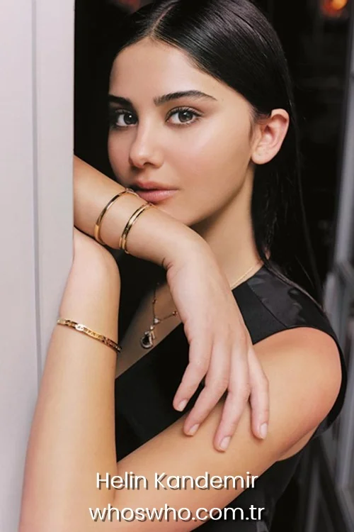 Close-up portrait of young Turkish actress Helin Kandemir with sleek straight dark hair, wearing a sleeveless black dress and accessorized with layered gold bracelets and a pendant necklace, posing by a doorway.
