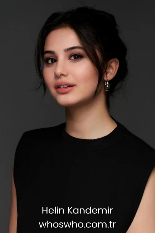 Close-up studio portrait of beautiful young Turkish actress Helin Kandemir with her dark hair tied back, smiling subtly and wearing a sleeveless black crewneck top and silver hoop earrings against a dark grey background.