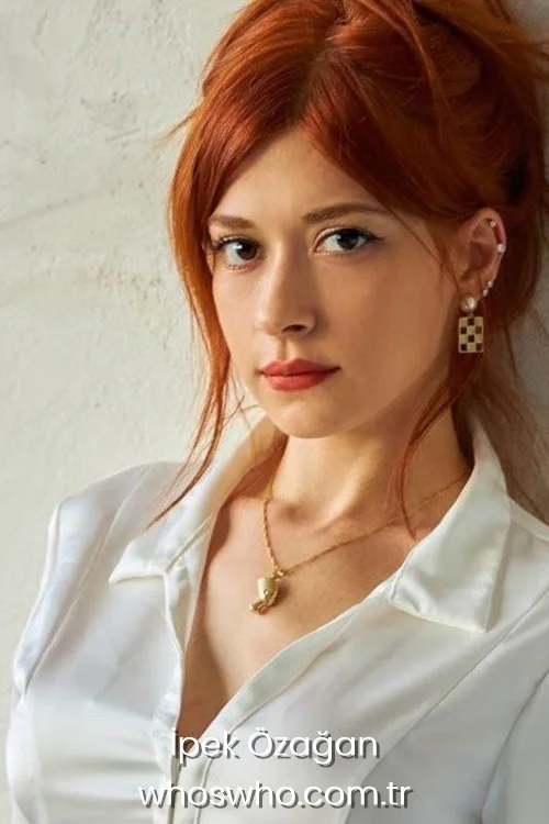 Turkish actress Ipek Ozagan with vibrant ginger-red hair tied back, wearing an elegant white satin collared shirt and a gold pendant necklace, leaning against a textured off-white wall.