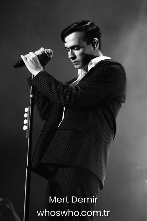 Mert Demir holding a microphone on stage during a black and white concert performance wearing a pinstripe suit.