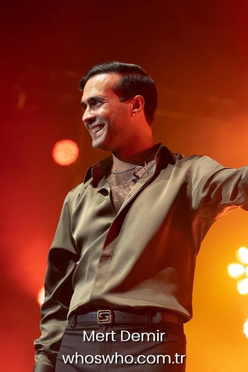 Mert Demir smiling on stage during a live concert wearing a khaki satin shirt.