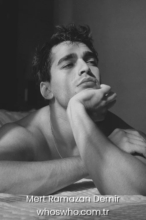 Black and white portrait of shirtless Mert Ramazan Demir lying on a bed with his hand on his chin. 