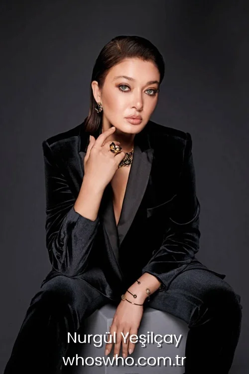 Studio portrait of Turkish actress Nurgul Yesilcay wearing a black velvet pantsuit and gold jewelry, sitting against a dark background.