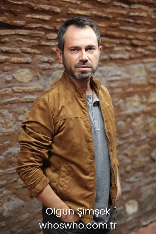Olgun Simsek in a brown suede jacket posing in front of a stone wall.