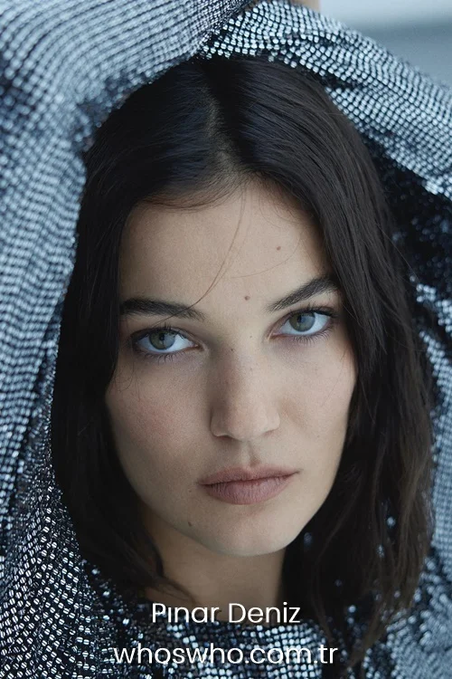 Turkish actress Pinar Deniz looking directly at the camera, framing her face with a sparkling silver textured fabric
