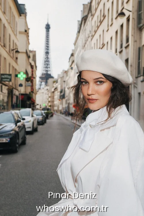 Turkish actress Pinar Deniz in Paris, wearing a white coat and a matching white beret with the Eiffel Tower in the background