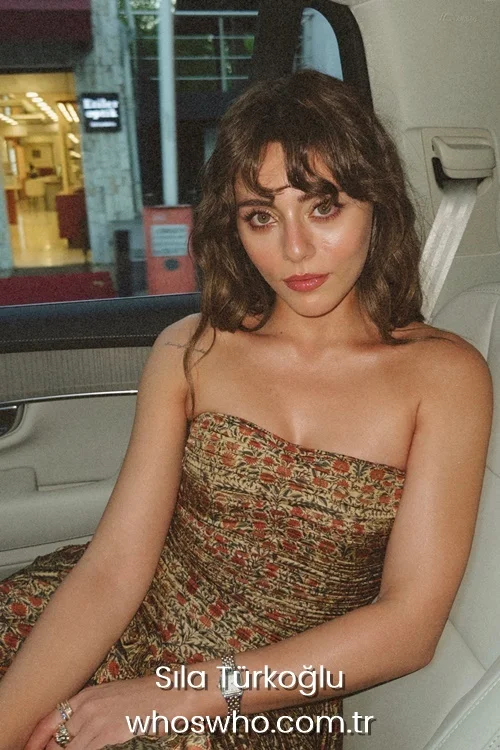 Turkish actress Sıla Türkoğlu with brown wavy hair and bangs wearing a strapless patterned dress inside a car.