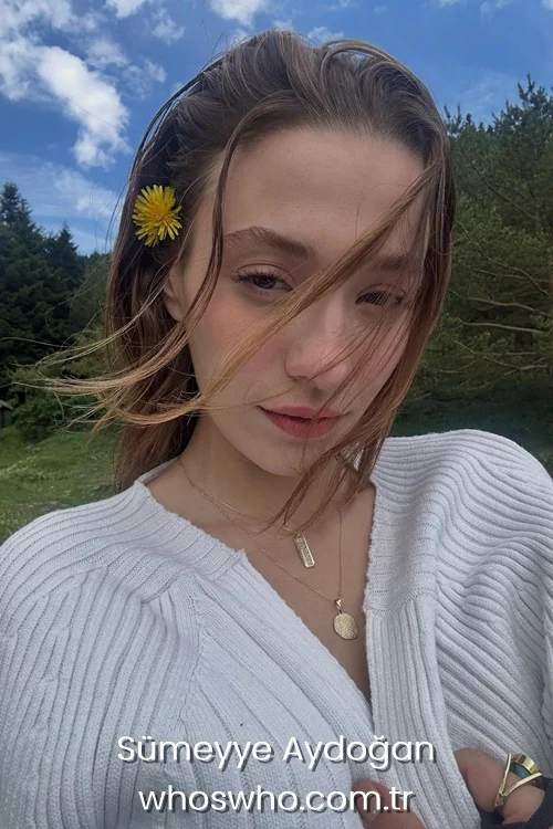 Turkish actress Sümeyye Aydoğan with a yellow dandelion flower in her hair wearing a white ribbed cardigan.