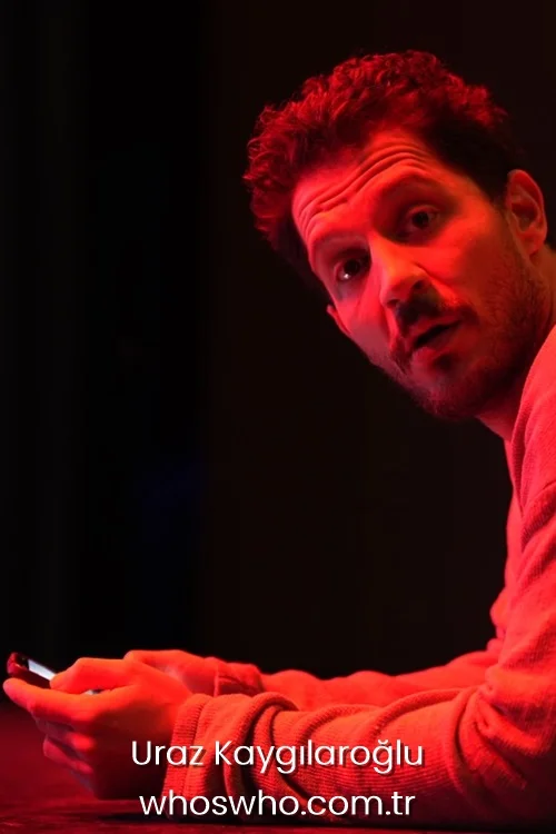 Turkish actor Uraz Kaygılaroğlu holding a smartphone under dramatic red stage lighting.
