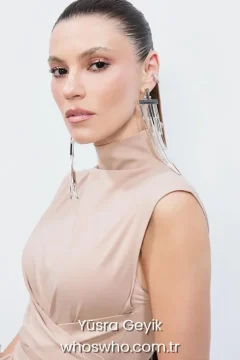 Turkish actress Yusra Geyik with a sleek high ponytail wearing a beige high neck dress and long silver statement earrings