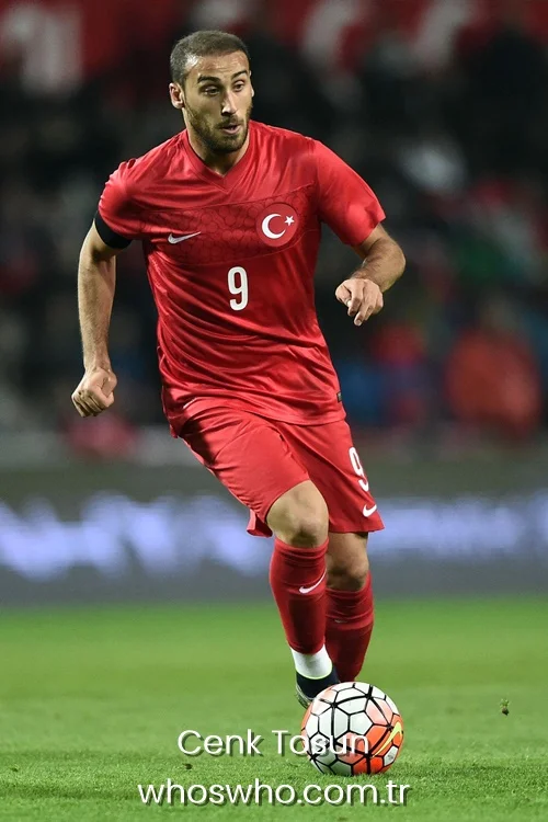 Turkish forward Cenk Tosun with a short buzz cut and beard, running and controlling a football on the pitch while wearing the red Turkey national football team jersey with number 9 and matching red shorts.