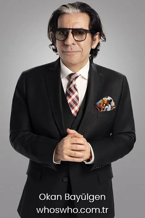 Famous Turkish talk show host Okan Bayulgen with dark hair and a signature white streak, wearing a black three-piece suit, a checkered tie, and glasses, standing with his hands clasped against a grey background.