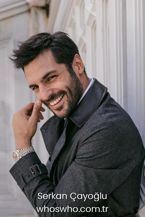 Serkan Cayoglu smiling while wearing a grey trench coat and a luxury watch.