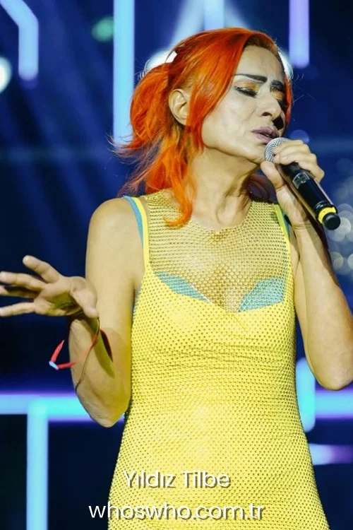 Yildiz Tilbe singing into a microphone on stage wearing a neon yellow mesh dress and bright orange hair.