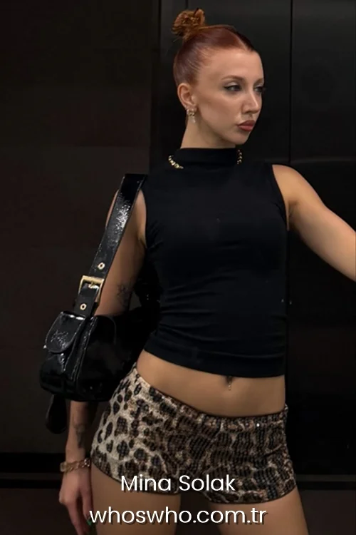 A fashion-focused portrait of Mina Solak posing with a high top knot bun hair style, wearing a sleeveless black turtleneck top and low-rise leopard print shorts, carrying a black patent leather shoulder bag against a dark background.