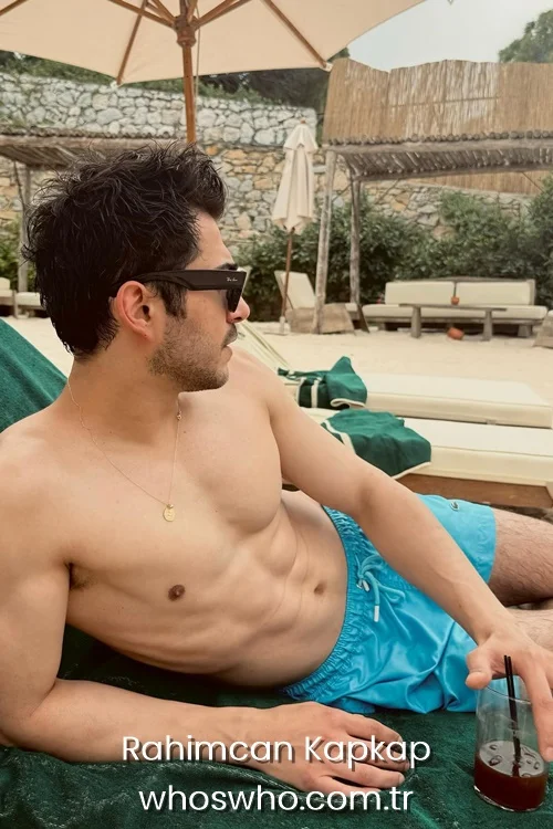Turkish actor Rahimcan Kapkap wearing blue swim shorts and sunglasses while relaxing on a beach sunbed.