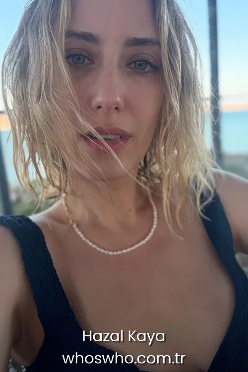 Close-up selfie of Turkish actress Hazal Kaya with blonde, wet-look wavy hair, wearing a black top and a thin necklace with the sea in the background.