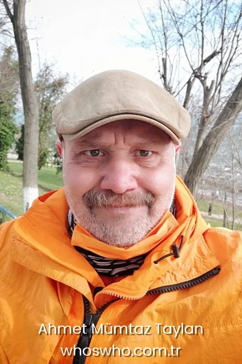 Ahmet Mumtaz Taylan smiling in an outdoor portrait wearing a beige flat cap and a bright orange jacket. 