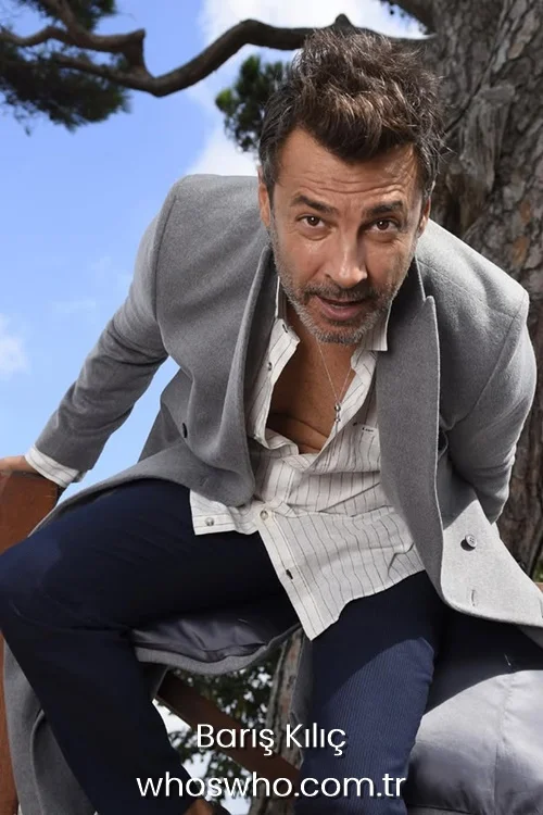 Baris Kilic leaning forward outdoors wearing a grey overcoat and a striped unbuttoned shirt.