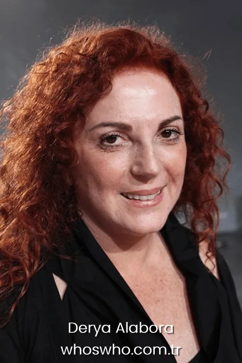 Close-up portrait of award-winning Turkish actress Derya Alabora smiling warmly at the camera, showcasing her signature long curly red hair and wearing a black sleeveless top.