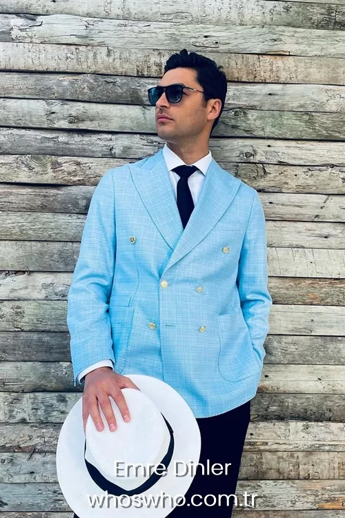 Emre Dinler in a light blue double breasted blazer jacket with gold buttons holding a white hat. 