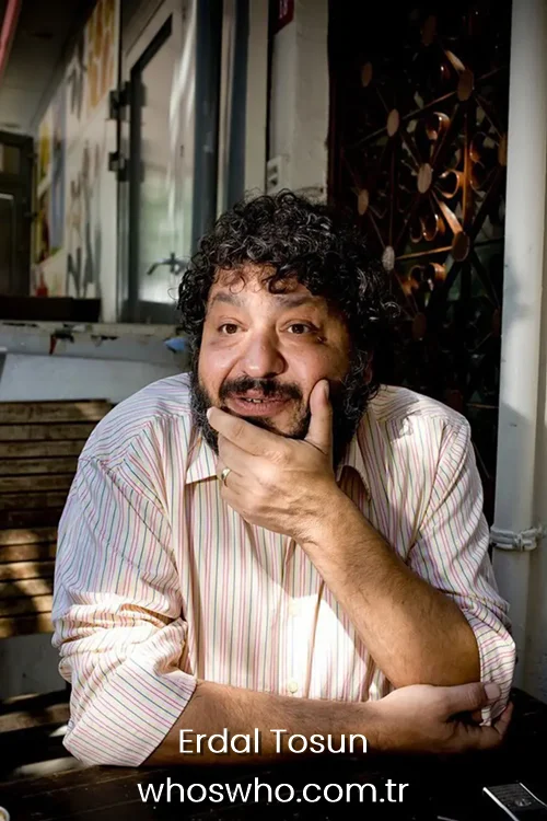 Turkish actor Erdal Tosun with a beard resting his chin on his hand at an outdoor table.