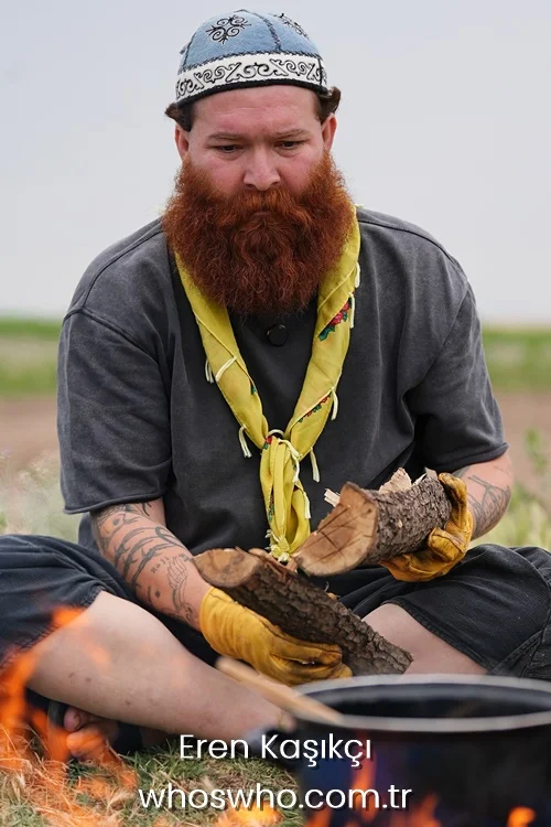 Turkish chef Eren Kasikci with a red beard wearing an embroidered skullcap and holding firewood next to a campfire