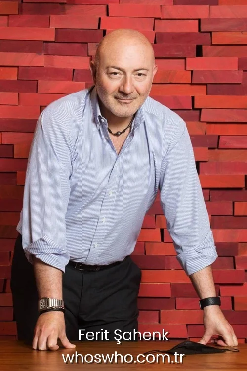 Turkish business executive Ferit Şahenk leaning on a table in a blue striped shirt against a red brick background.