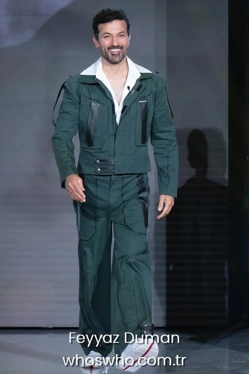 Turkish actor Feyyaz Duman smiling and walking on a fashion runway wearing a green utility suit with a white collar shirt