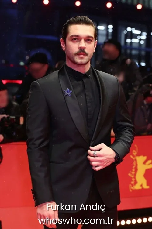 Turkish actor Furkan Andic posing on the Berlinale red carpet, with slicked-back hair and a mustache, wearing a sharp black tuxedo jacket, black button-down shirt, and a blue flower lapel pin.