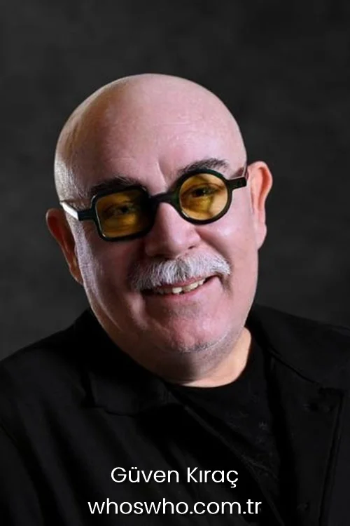 Close-up portrait of Turkish actor Güven Kıraç smiling broadly against a dark grey background, wearing a black shirt and distinctive black-framed glasses with yellow tinted lenses.