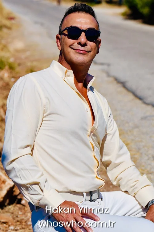 Medium shot of Turkish actor Hakan Yılmaz sitting outdoors on a sunny day, smiling confidently while wearing a light cream long-sleeved button-up shirt, white trousers with a silver belt buckle, and stylish black sunglasses.
